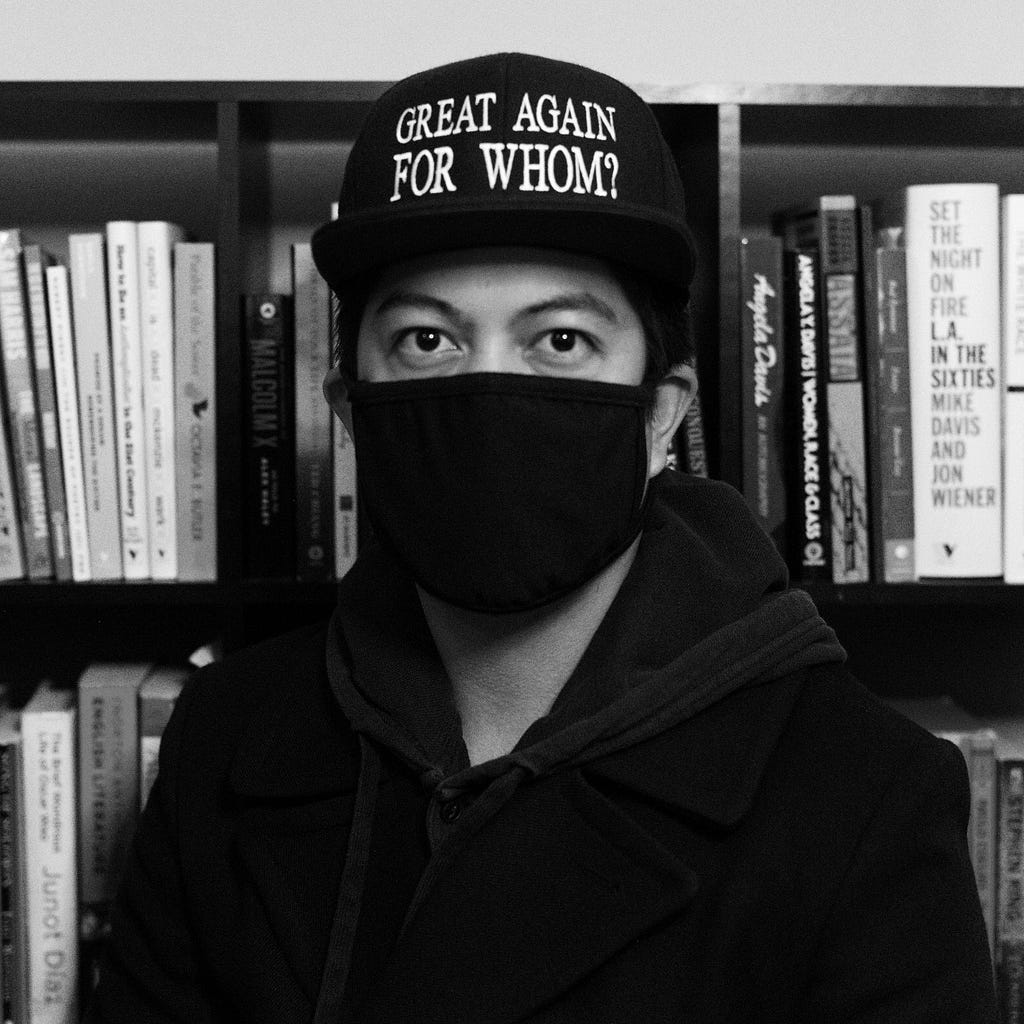 This is an image of the poet, Michael Guevarra. He is masked, wearing all black and a hat that reads “Great Again For Whom?”