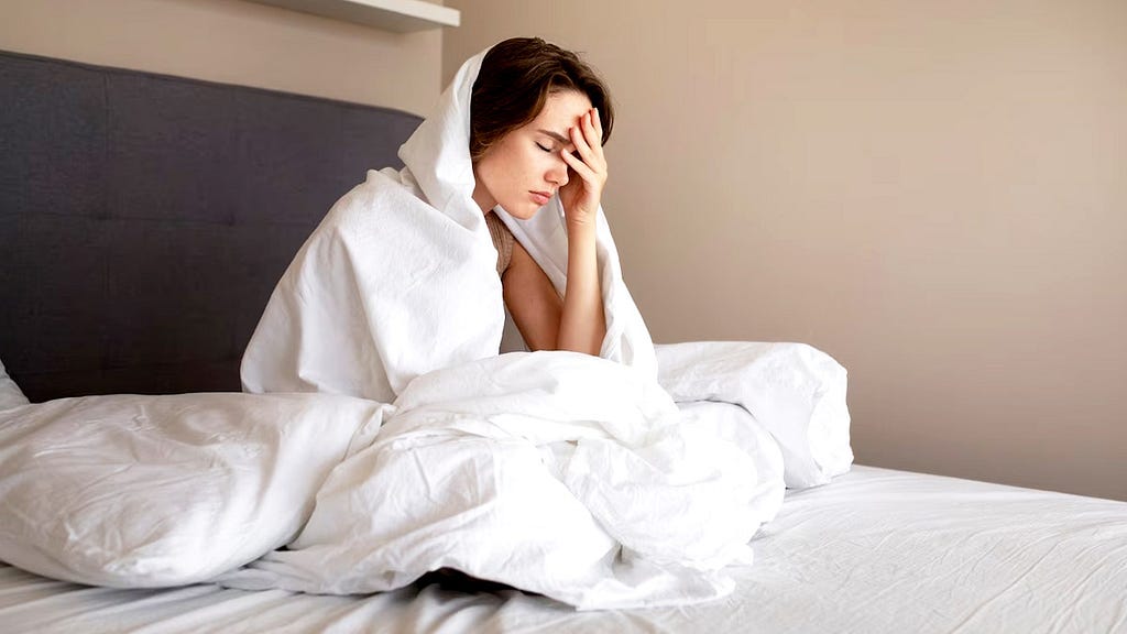 Woman in bed wrapped in bed sheets and in a bad mood. She looks tired.