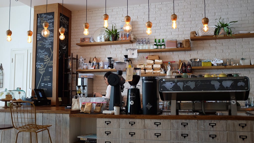 A welcoming, simple and clean, independent coffee shop. A barista is seen making a drink behind the bar.