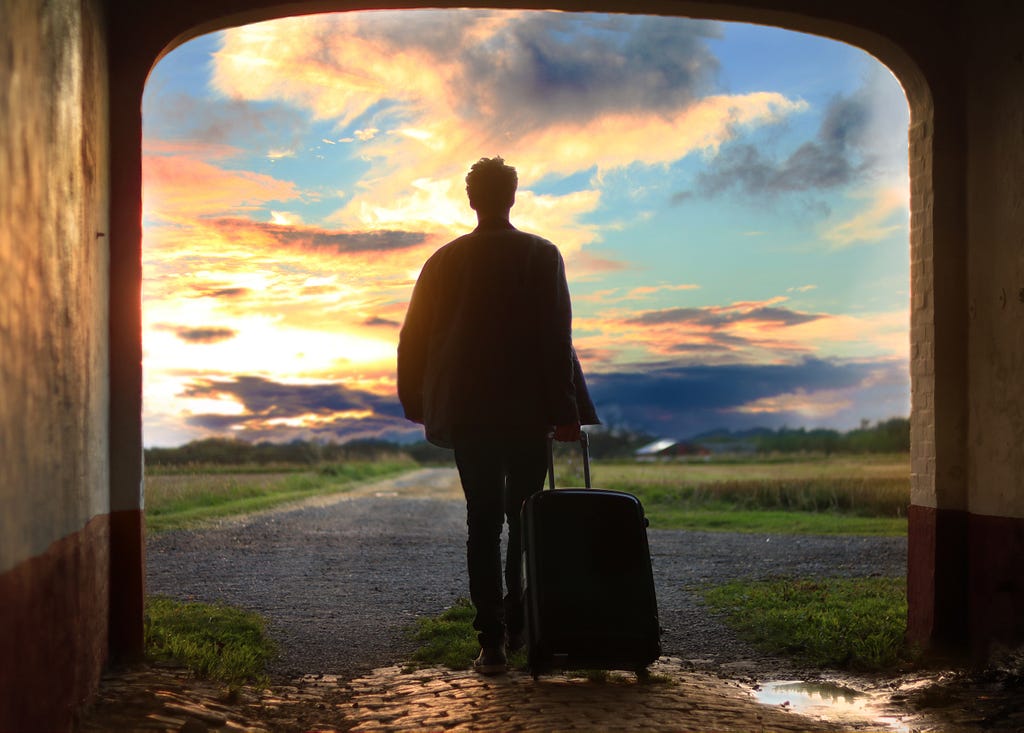 Man with a suitcase walking toward a cloudy sunset.