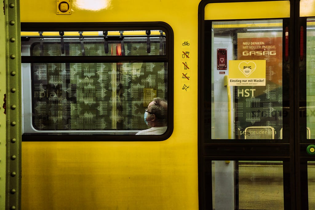 A yellow train from the outside with a sign showing that it is only allowed to enter with a mask. A man sits inside the train, wearing a mask.