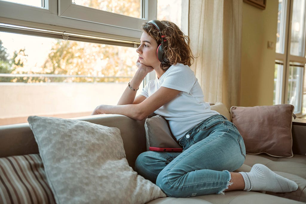 A photograph of a young person kneeling on a couch and leaning over the back of it to stare out the window. They have headphones over their ears.