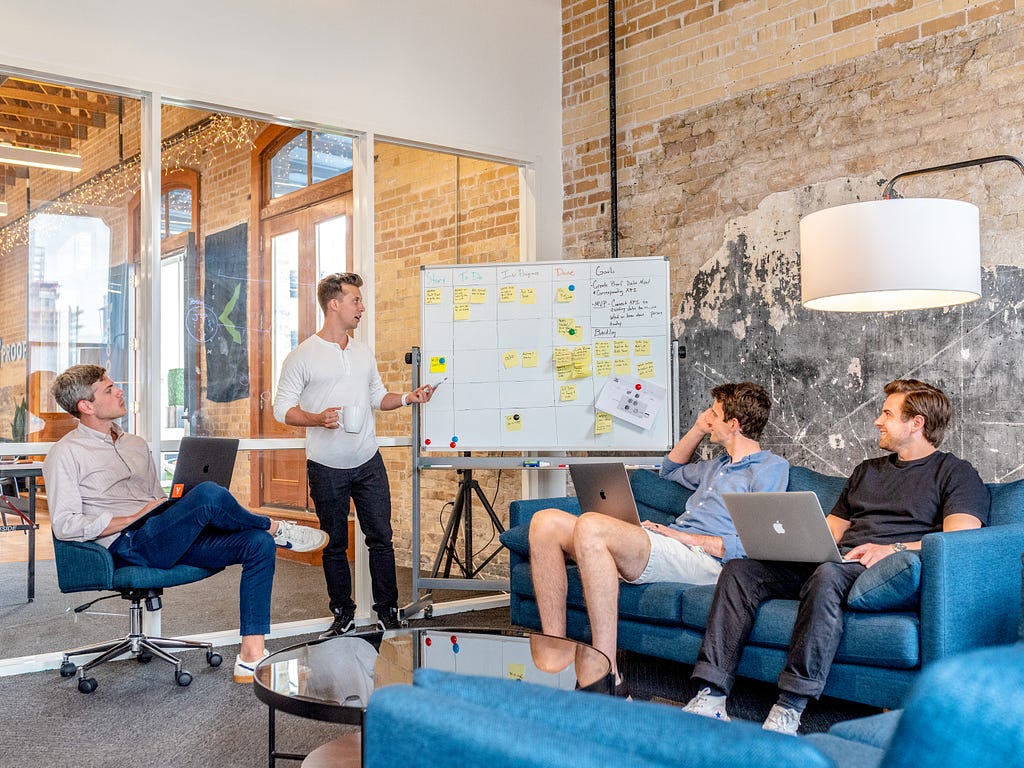 Brainstorming session of a team of four men. Three of them sitting with laptops on each’s lap and one making analysis using a sticky notes on white board.