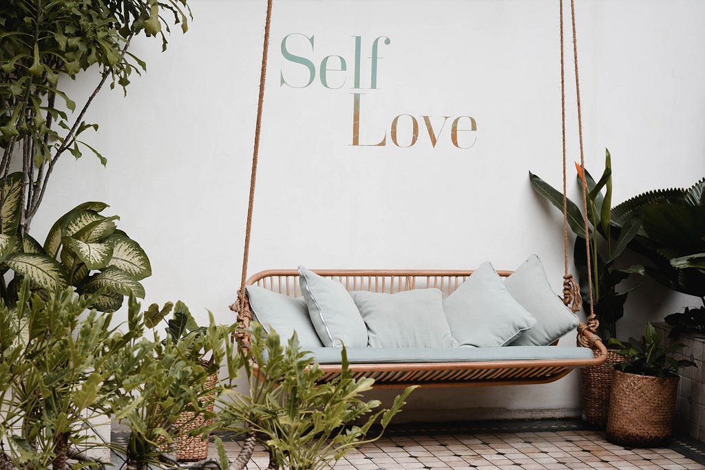 A scene of comfort with a rustic style couch, plants, and a white wall with the phrase ‘self love’ on it.