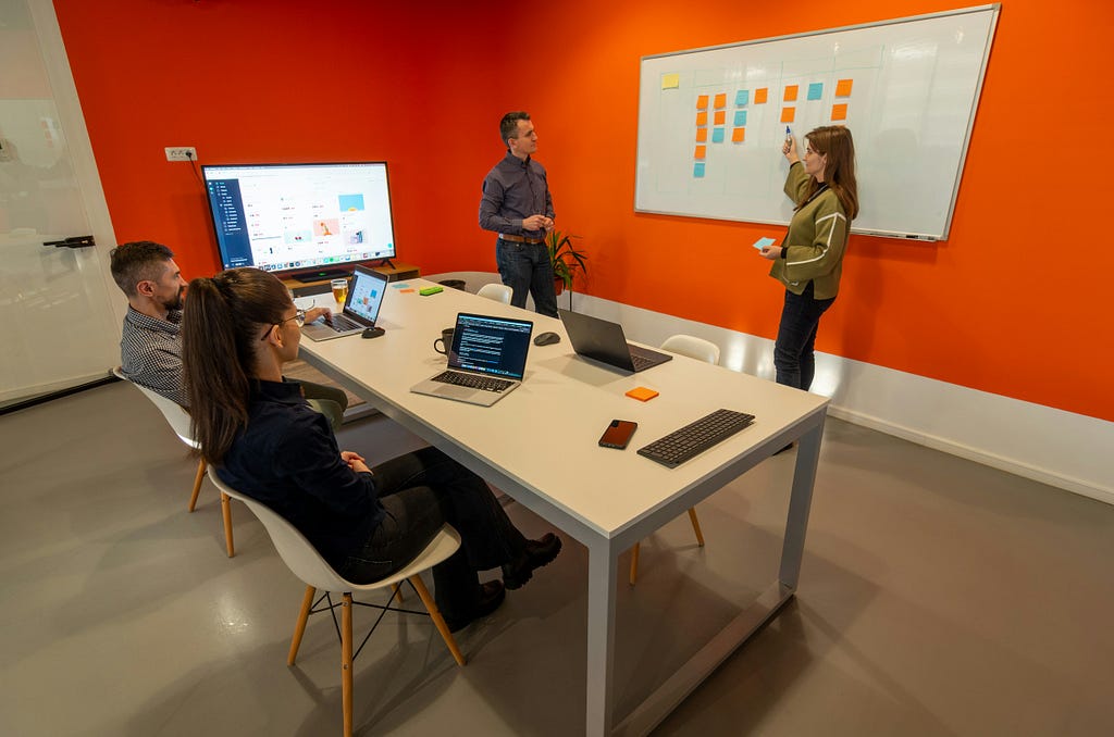 A meeting room with people discussing a project. Two people sit at a table next to a large monitor screen while two people present a whiteboard with sticky notes.