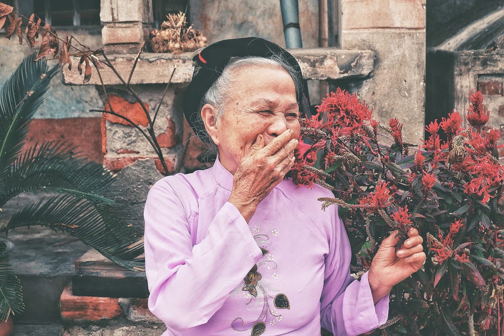 An elderly woman in a purple outfit, smiling while next to a bush of red flowers.