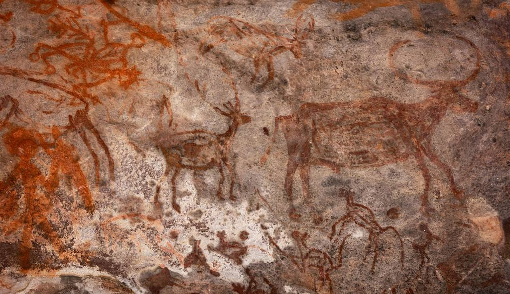 Ancient cave painting depicting various animals, including deer and large-horned cattle, alongside human figures engaged in hunting scenes. The artwork is created using reddish-brown pigments on a rough, textured rock surface. The composition includes dynamic movement, with hunters wielding spears and bows, and animals in different postures. The style reflects early human storytelling and artistic expression, likely from prehistoric times.