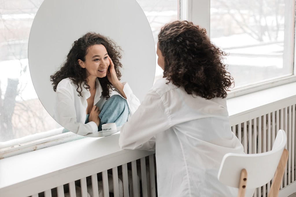 a woman in a white top smiles at herself in the mirror after doing her self-pleasure practice