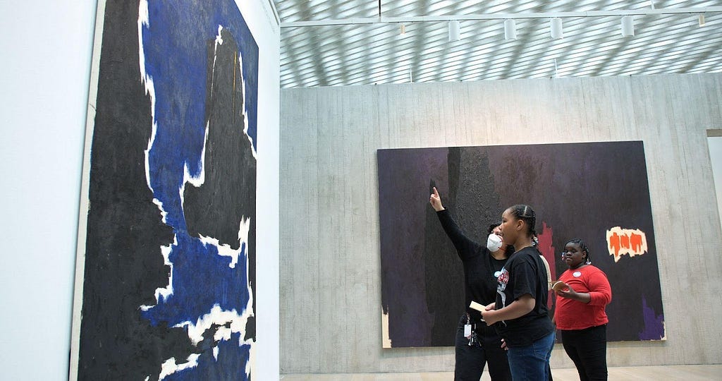 An educator points at a massive painting in a gallery with two young students who hold sketchbooks and look at the painting.