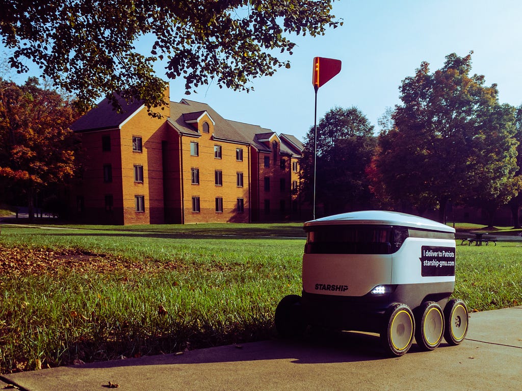 A delivery robot on a sidewalk is seen in the foreground with a building and a grassy land in the background