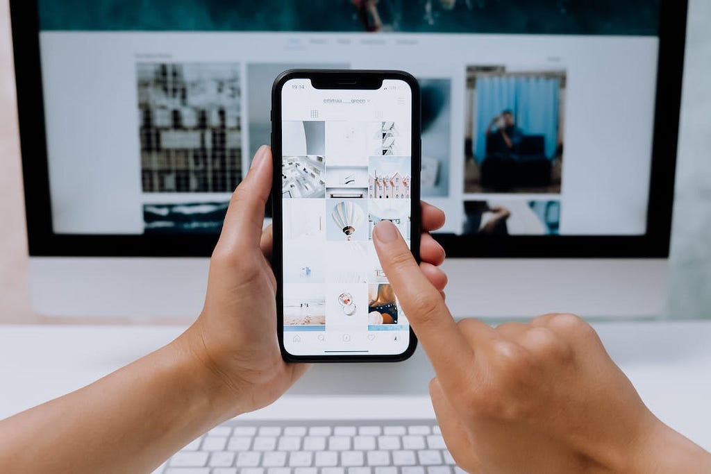 A cropped photo of a person’s hands scrolling through stock photos on a phone while working on a PC. cottonbro/Pexels.