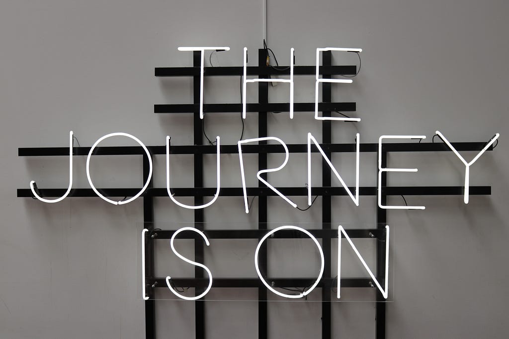 A photograph of a white-light neon sign with the words “The Journey Is On” set agains a black grid hanging on a gray wall. Photo by Clemens van Lay on Unsplash.