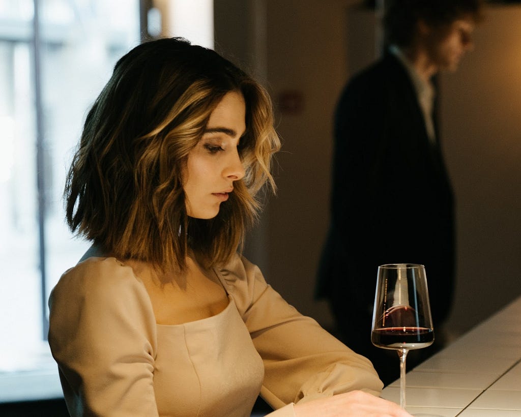 woman looking at a glass of wine as her date stands in the distance