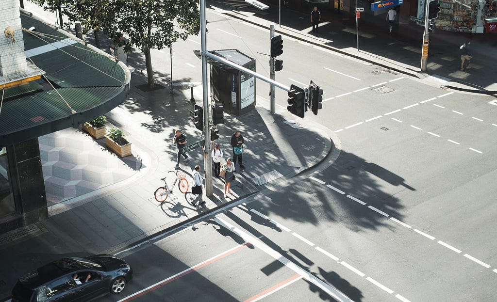 Image of a bright street with a car and bicycle lanes, few people are waiting on the traffic light to cross the street, no traffic at all, just another sunny morning in a quite cross road.