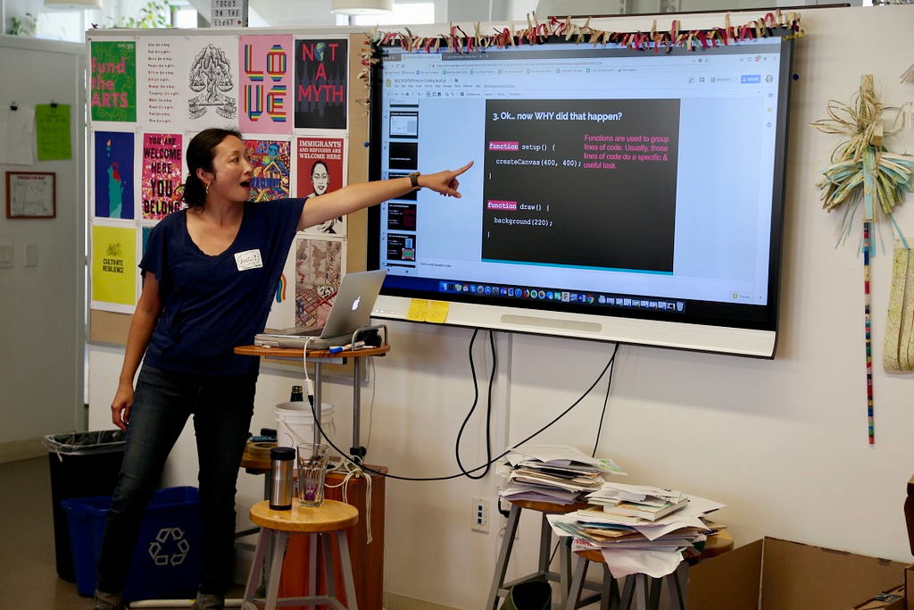 Photograph of Angi standing by a screen with the projection of a presentation on it. She points at the screen and is speaking. The screen says, “Ok… now WHY did that happen?”