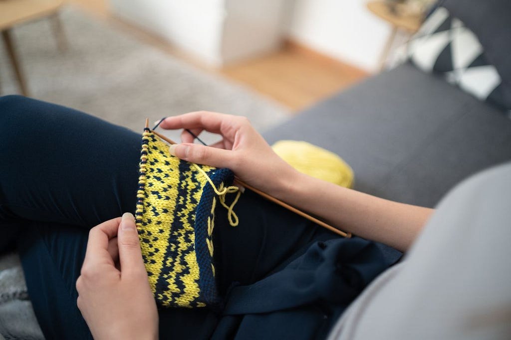 Legs and torso of a person sitting on a couch holding yellow and blue knitting in their hands