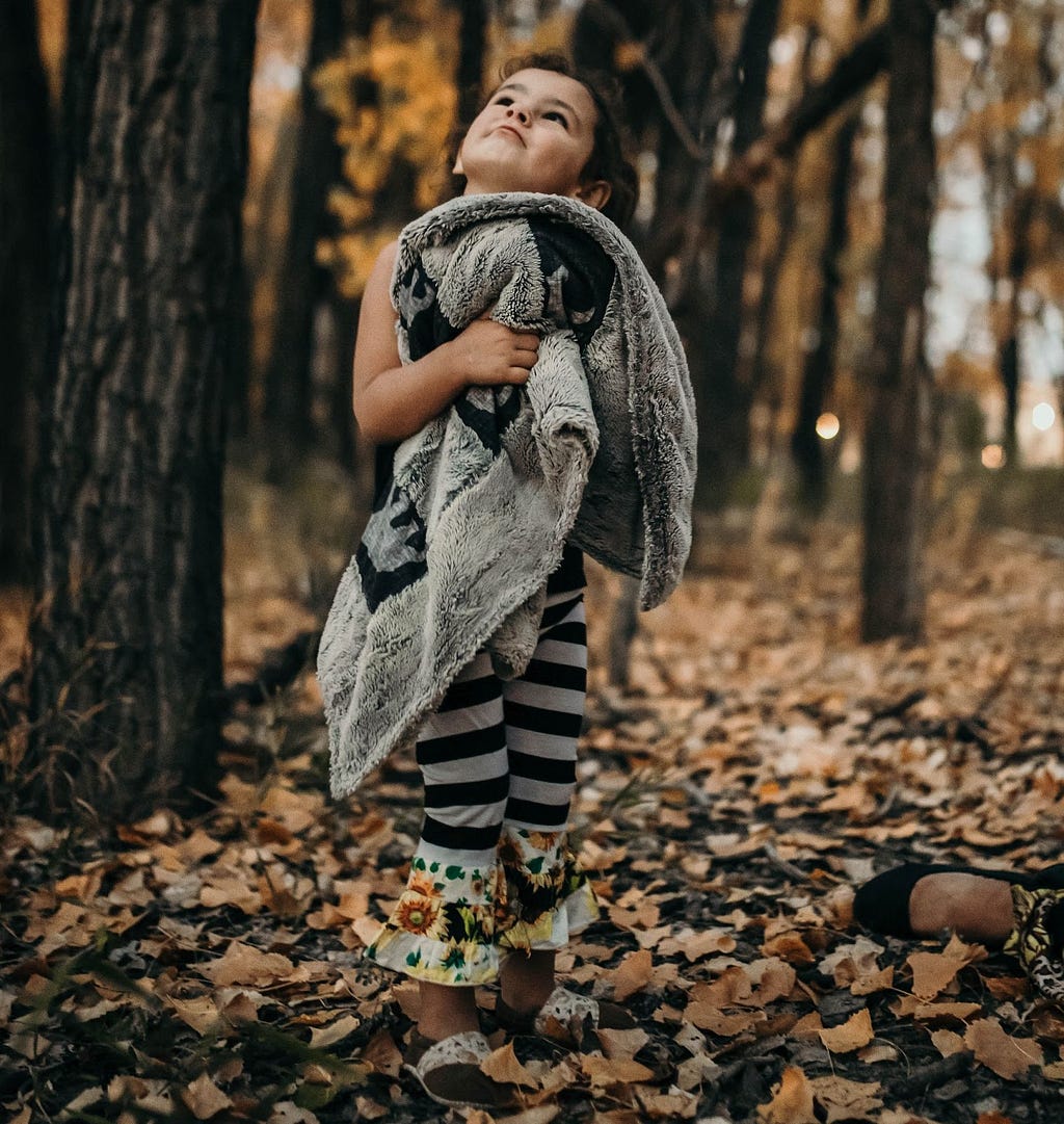 Young toddler holds blanket outside and looks up at the trees