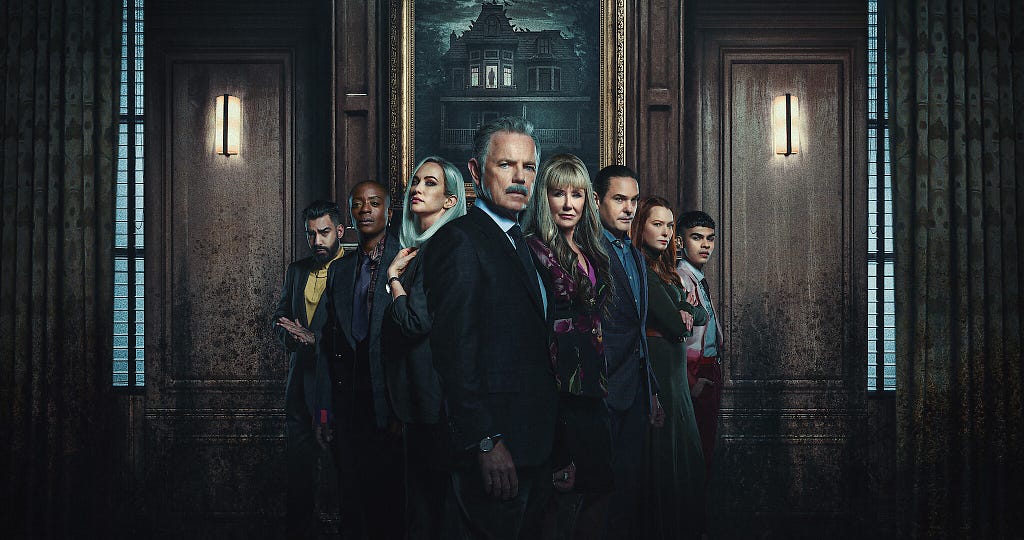 An image shows the main cast of The Fall of the House of Usher standing in a triangle formation. From far left to far right are Rahul Kohli, T’Nia Miller, Kate Siegal, Bruce Greenwood, Mary McDonnell, Henry Thomas, Samantha Sloyan, and Sautiyan Sapkota. The cast stands in a sombre room with the portrait of an old bungalow behind them.