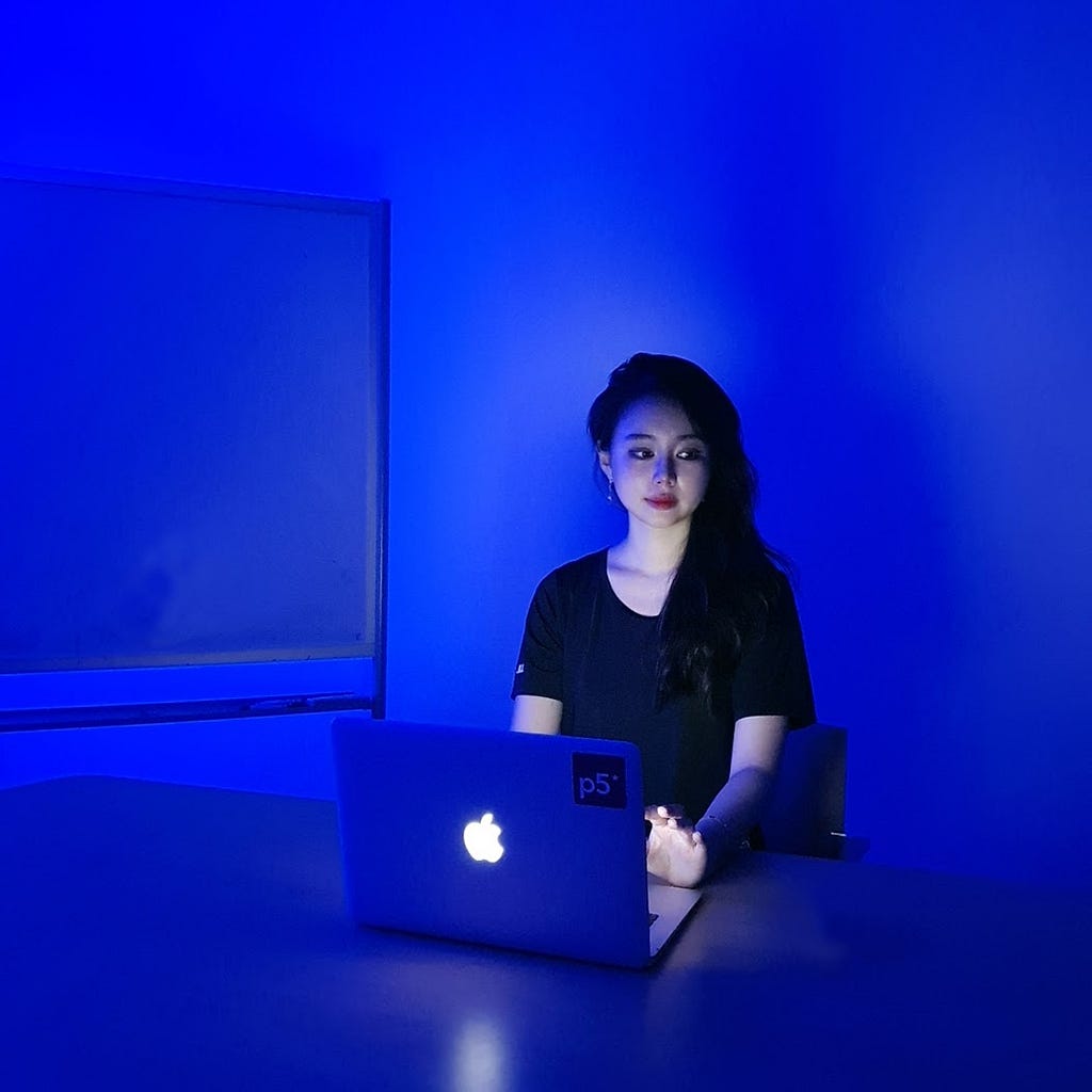 Inhwa with long hair, wearing a T-shirt, is seated in front of a laptop at a desk. She looks to her left.