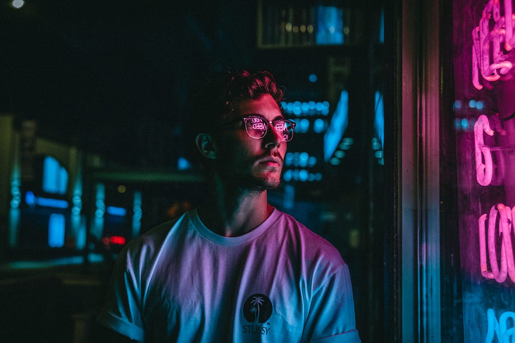 Man in white t-shirt walking in a city during the nightime, stopping to look at a pink neon sign in a window, the pink glow reflecting off his sunglasses.