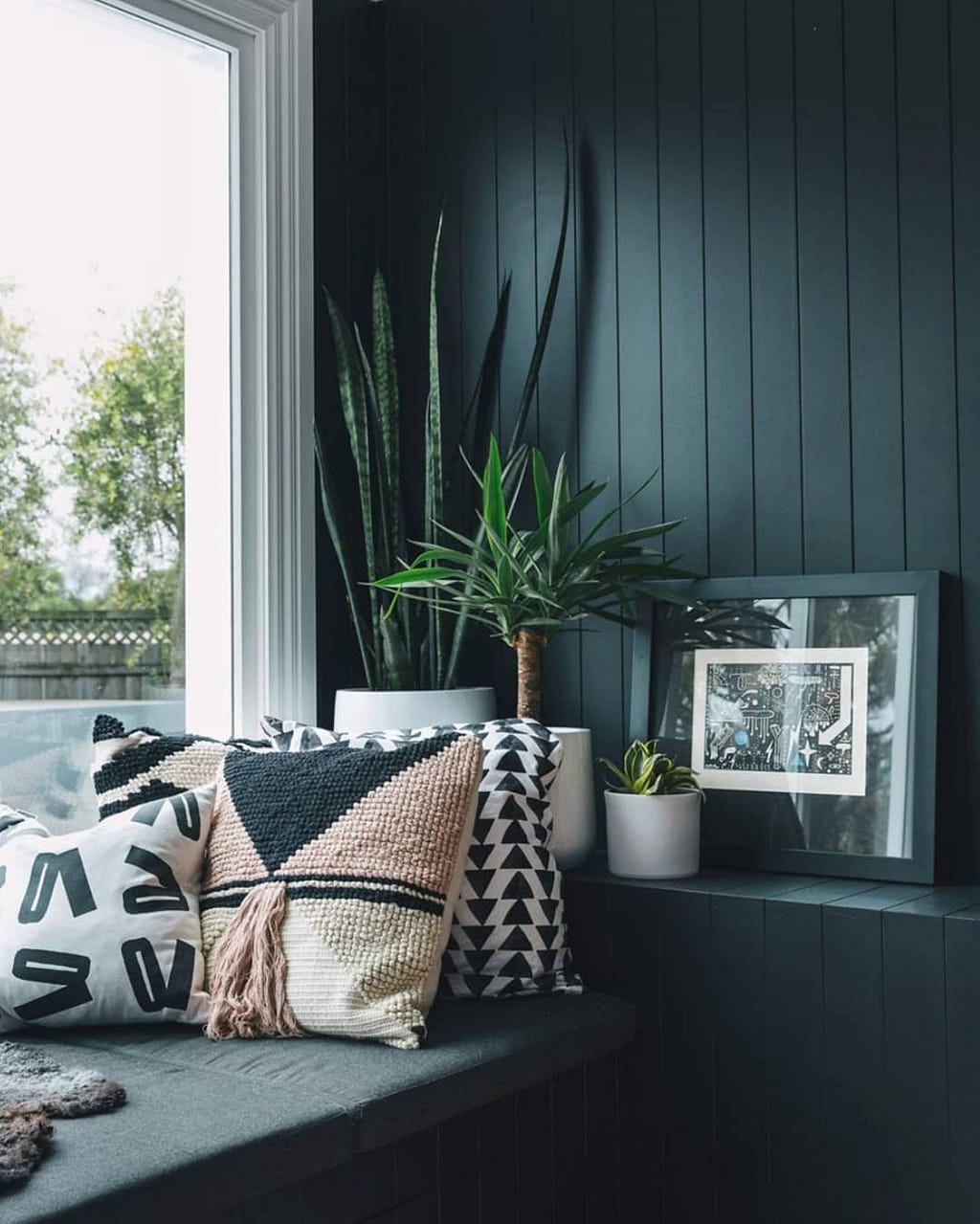 A corner of the room by the window with a black wall and bench, decorative pillows, and house plants.