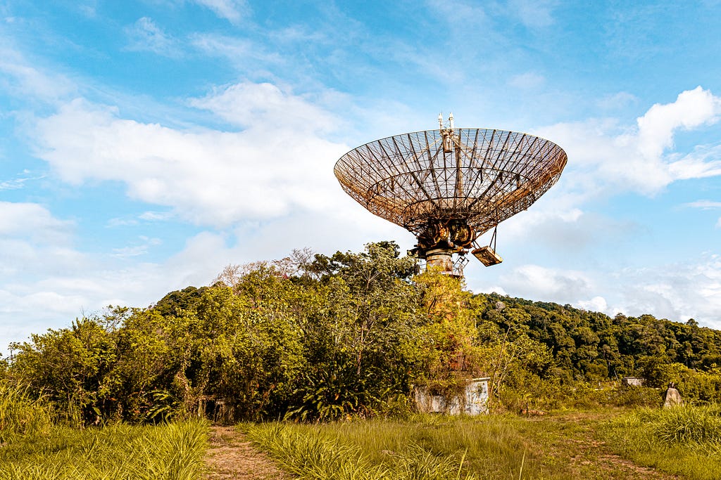 Foto de uma construção baixa abandonada, tomada pela natureza e com algumas árvores ao redor. Sobre a construção existe uma grande antena parabólica apontada para o céu. Ao fundo, temos um céu azul com nuvens brancas.