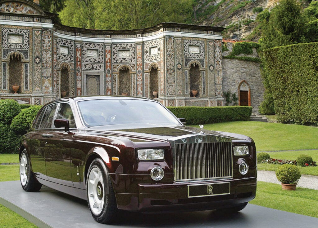 Un Rolls-Royce Phantom de 2003, creado bajo BMW, está estacionado frente a un edificio ornamentado. El coche tiene una parrilla distintiva, faros redondos y rectangulares, y puertas traseras suicidas.