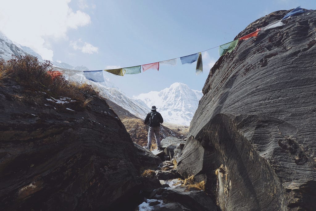 A person standing between two rocks staring at the mountains. Tibetan prayer flags are hung over their head.