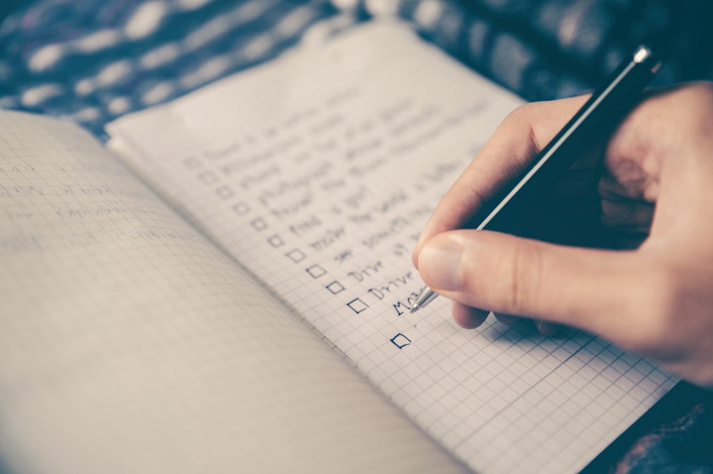 Uma pessoa escrevendo com uma caneta em um caderno pequeno, compilando sua lista de atividades.