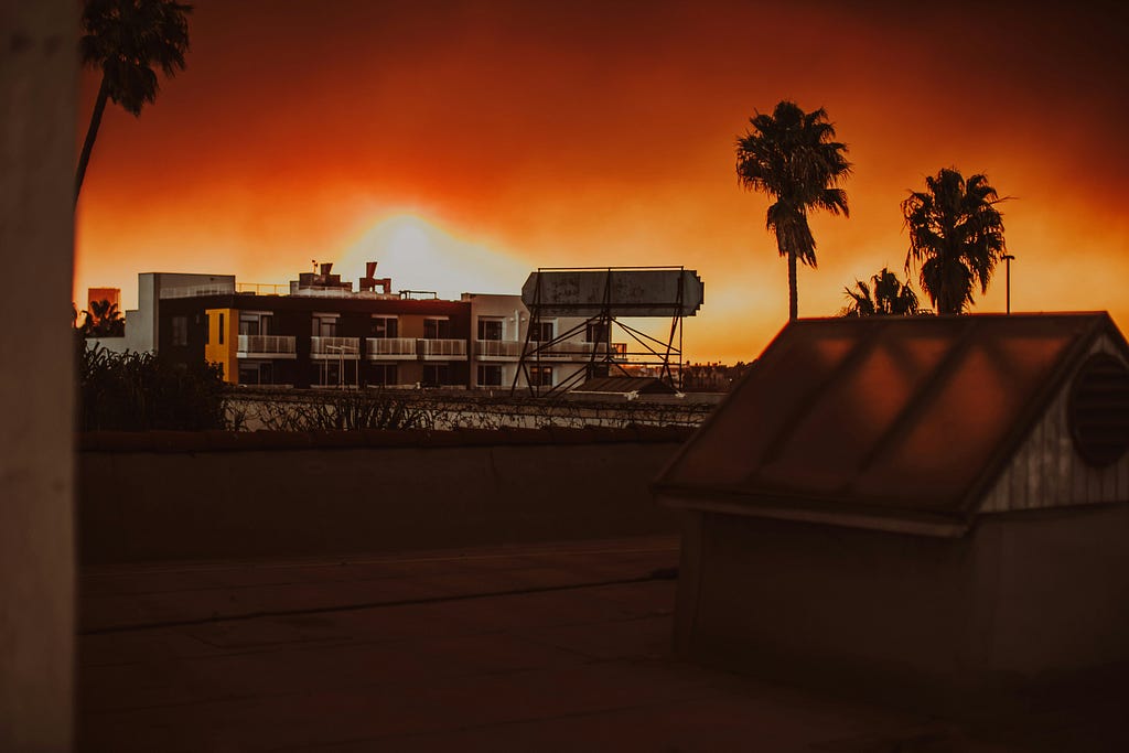 A view of two buildings with palm trees in the background. The sky is dark and filled with smoke, an orange from a nearby fire on the horizon illuminates the scene.