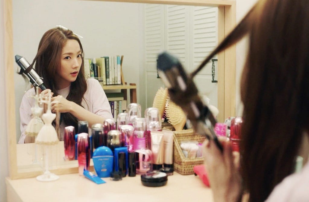 Kim Mi-so getting ready for work, hair curler in hand, sitting in front of a dressing table full of cosmetics.