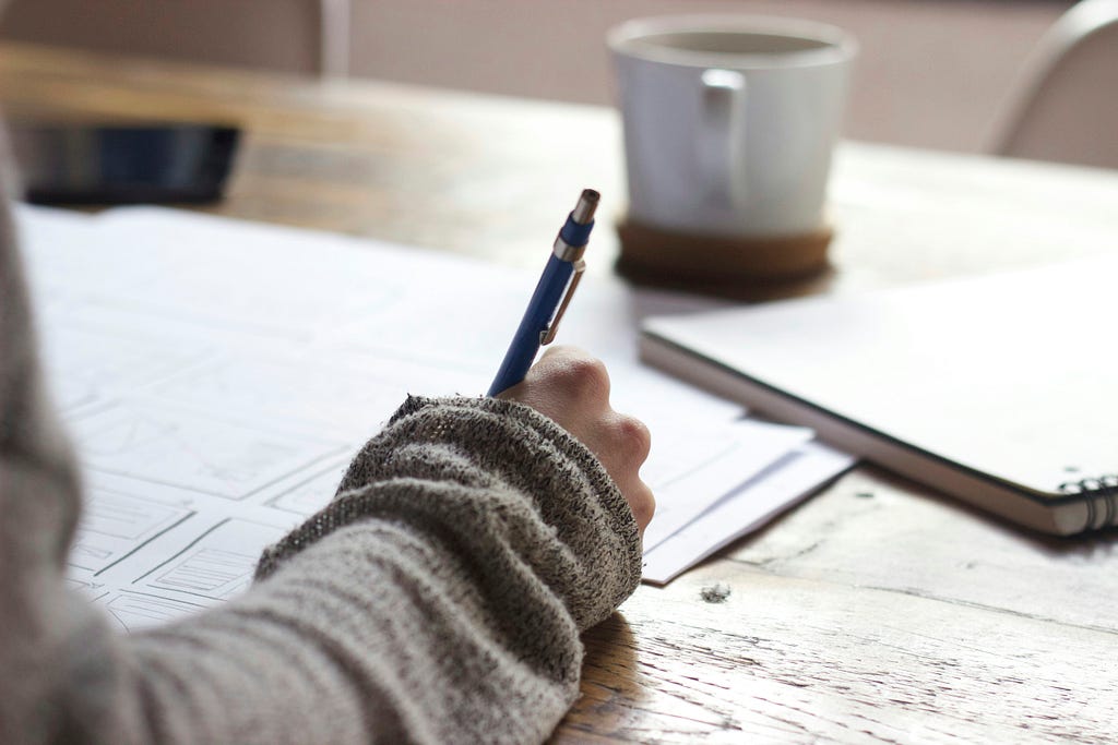 This image depicts a person in a sweatshirt writing something on a piece of paper with a pen, a notebook, and a cup of coffee on top of a table.