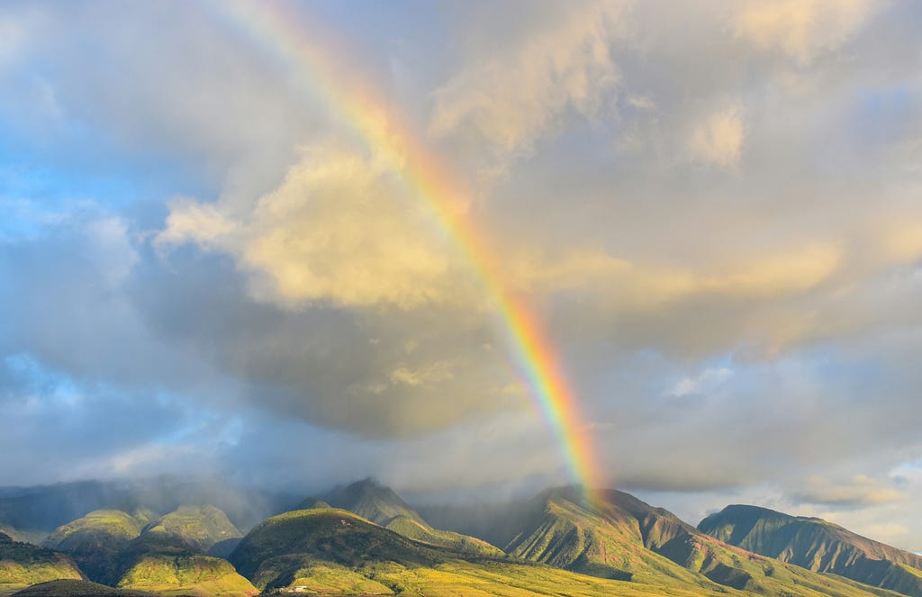A segment of a rainbow reaching from sky to mountain in a range, with a backdrop of sky and painterly clouds.