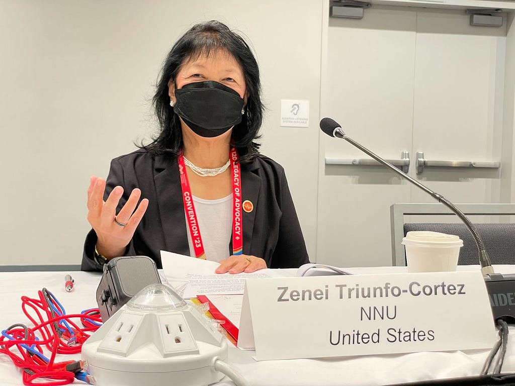 Filipina woman wearing black mask sitting at table with microphone in front and holding hand palm up, sign in front reads Zenei Triunfo-Cortez, NNU, United States.