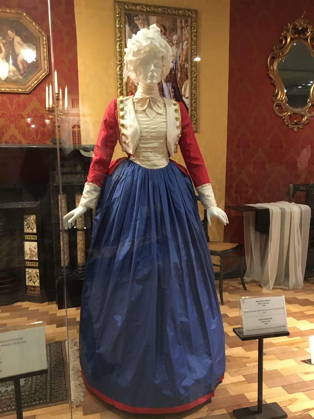 Um manequim feminino usando uma réplica de um vestido da Era Napoleônica, com um casaco vermelho com detalhes brancos em estilo militar da época, parte superior do vestido branca, uma saia azul escura, longa, e uma peruca cacheada branca.