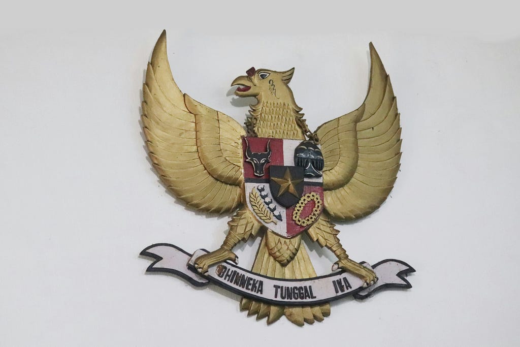 A photo of an Indonesian Garuda plaque mounted on a white wall. The plaque symbolises the Indonesian Pancasila and the text “Bhinneka Tunggal Ika”