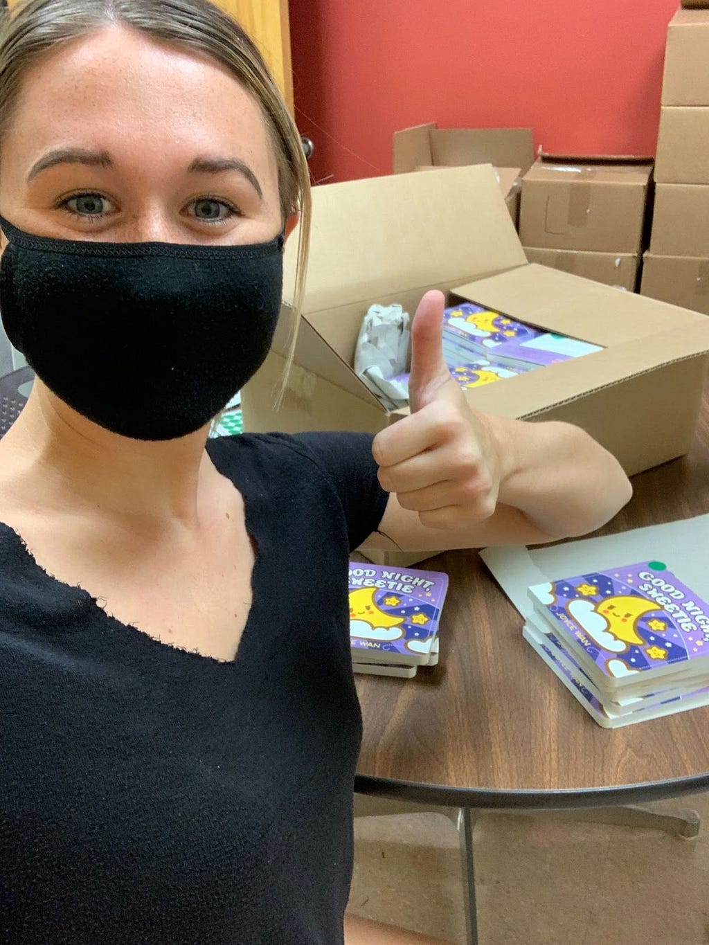 A masked woman gives a thumbs up in front of boxes she is packing with children’s books.