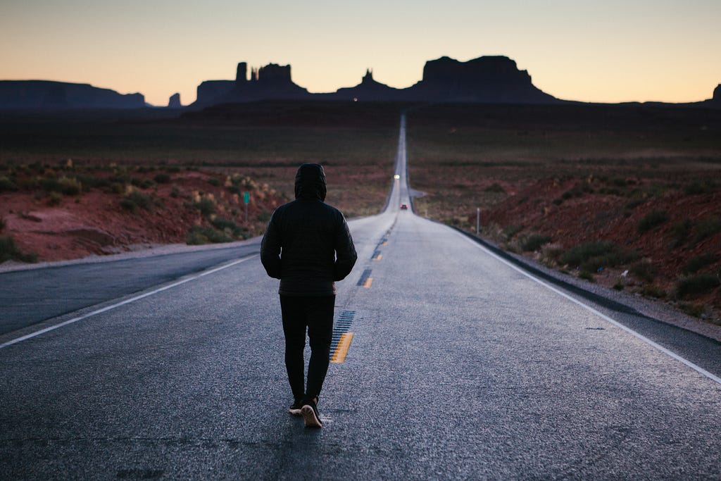 A man in a hoodie walking away from the camera along a long, straight road in the desert