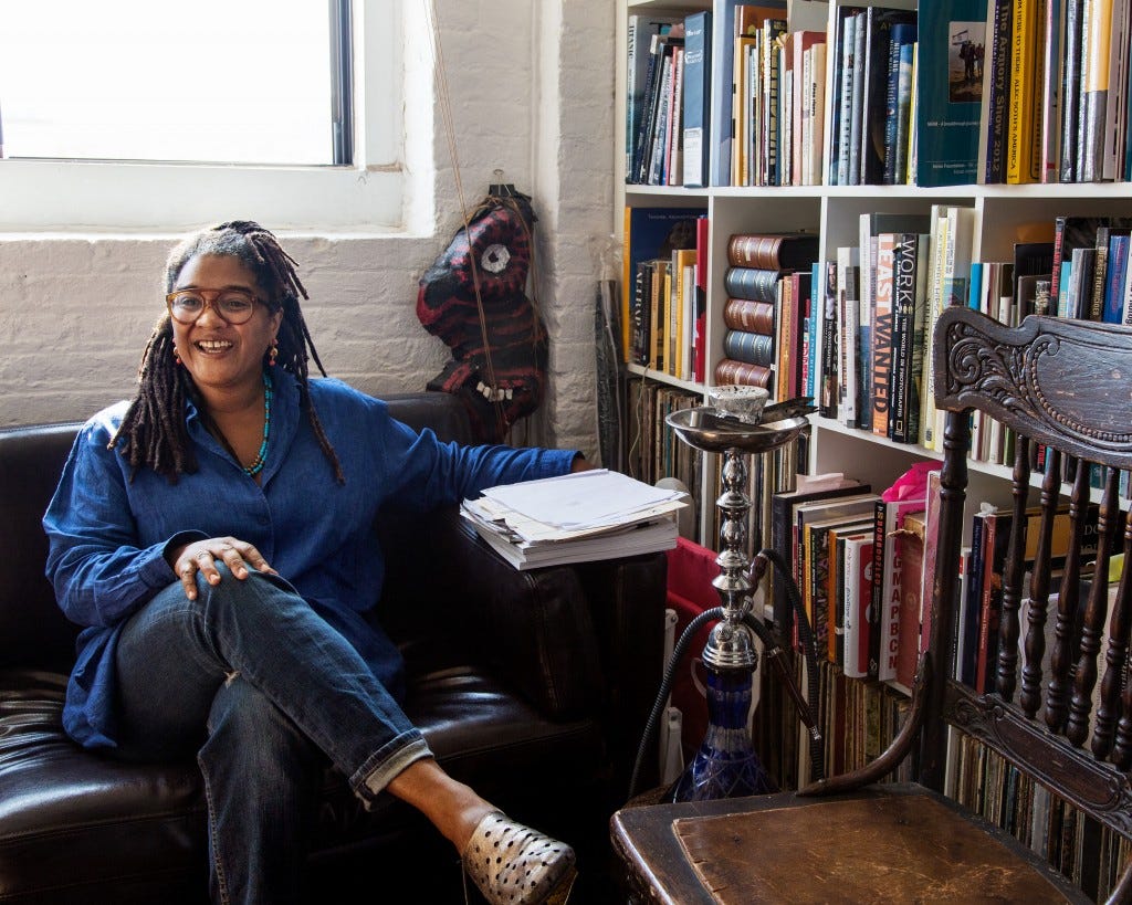 A woman with brown skin and brown hair in dreadlocks wearing a blue shirt and jeans sits on a black couch. A pile of papers is to her right, along with statues and a wall of bookshelves.