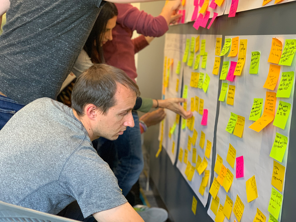 An image of a man in a gray shirt in the foreground looking at a wall with white background that has post-its with handwriting