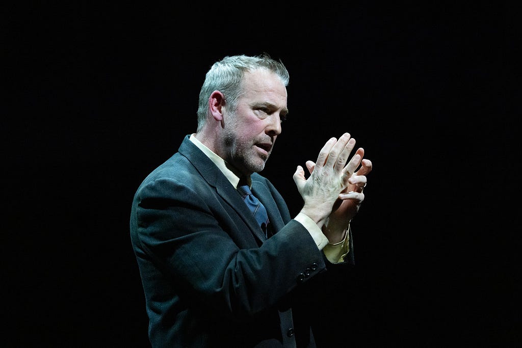 A man in a gray suit holds his hands in front of him as if catching or holding something.