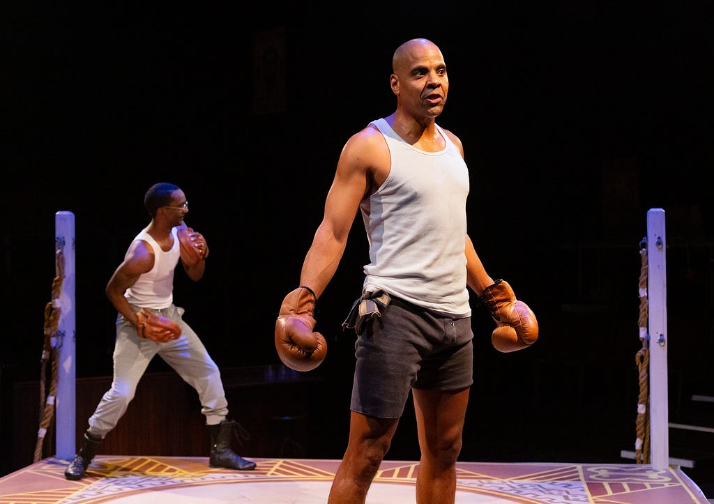 A man with brown skin and short hair boxes in the background in a tank top while another man with brown skin and a shaved head talks in the foreground, wearing a white tank top and boxing gloves.