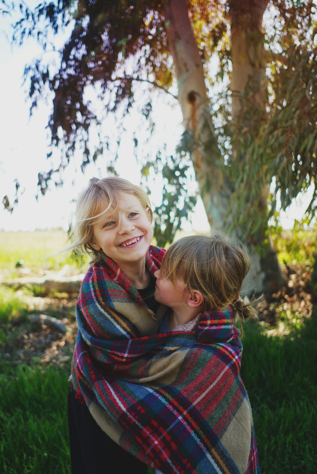 The image is of two smiling young girls, standing upright, wrapped in a blue, red, and green Scottish-checked blanket. The taller, a blond faces the camera, while the younger, shorter, dark ponytail faces her sister. They are outside. The tree in the background is a eucalyptus. It is a sunny day.