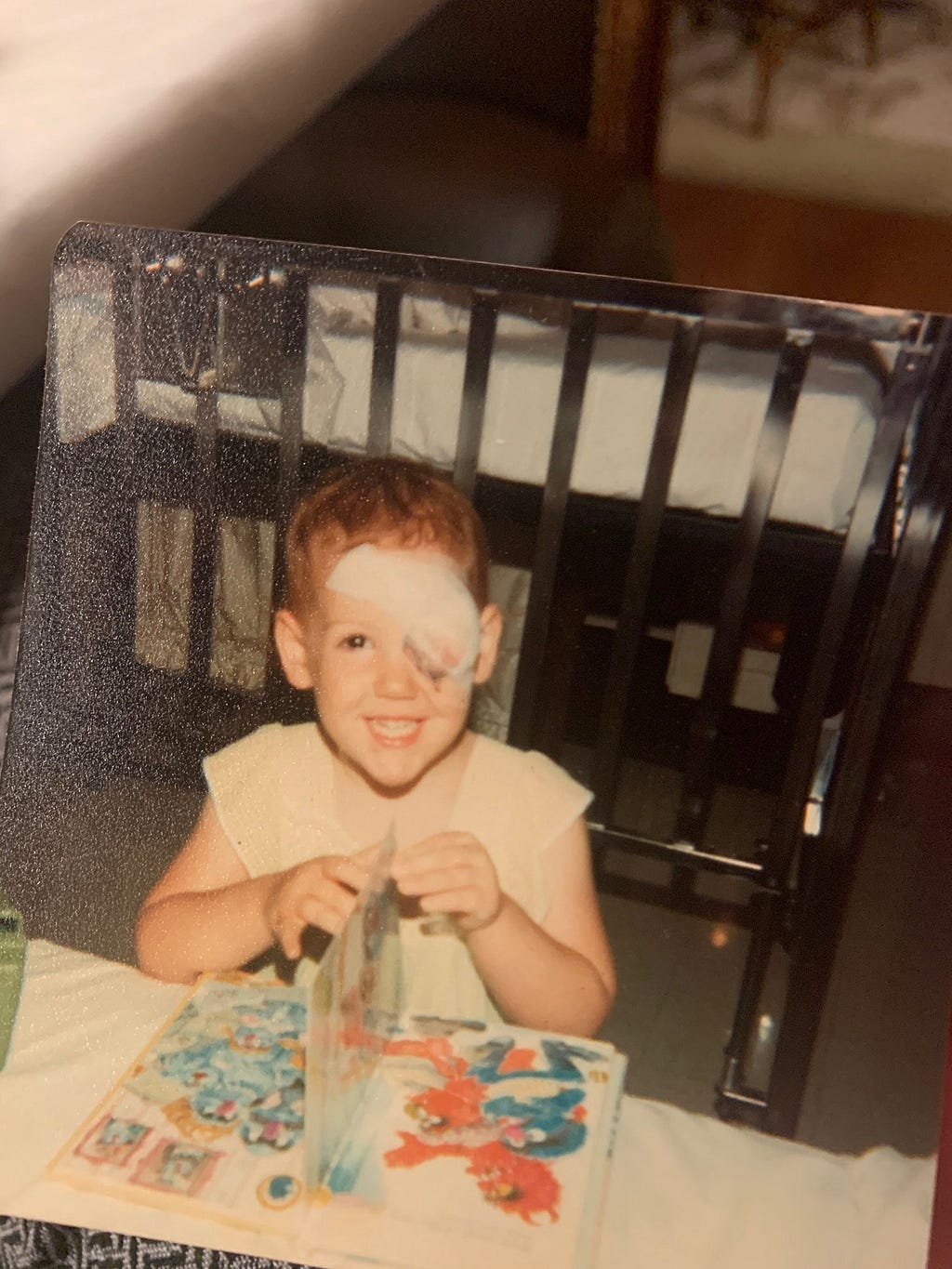 Young child with blood-stained patch over left eye looking at Sesame Street book and smiling at the camera. A hospital bed is visible in the background.