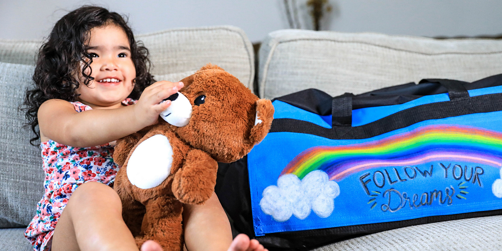 A smiling child sits on a couch holding a teddy bear. Next to her is a duffel bag decorated with the phrase, “Follow your dreams.”