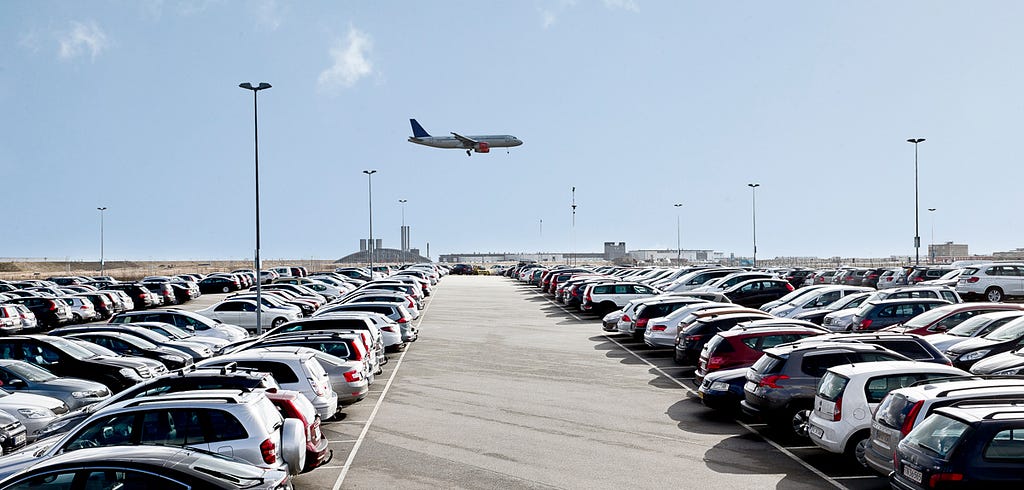 An airport car park with aeroplane flying overhead