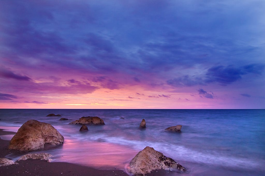 A beach with boulders on the shore shows a pinkish orange sunset surrounded by bluish, gray clouds.