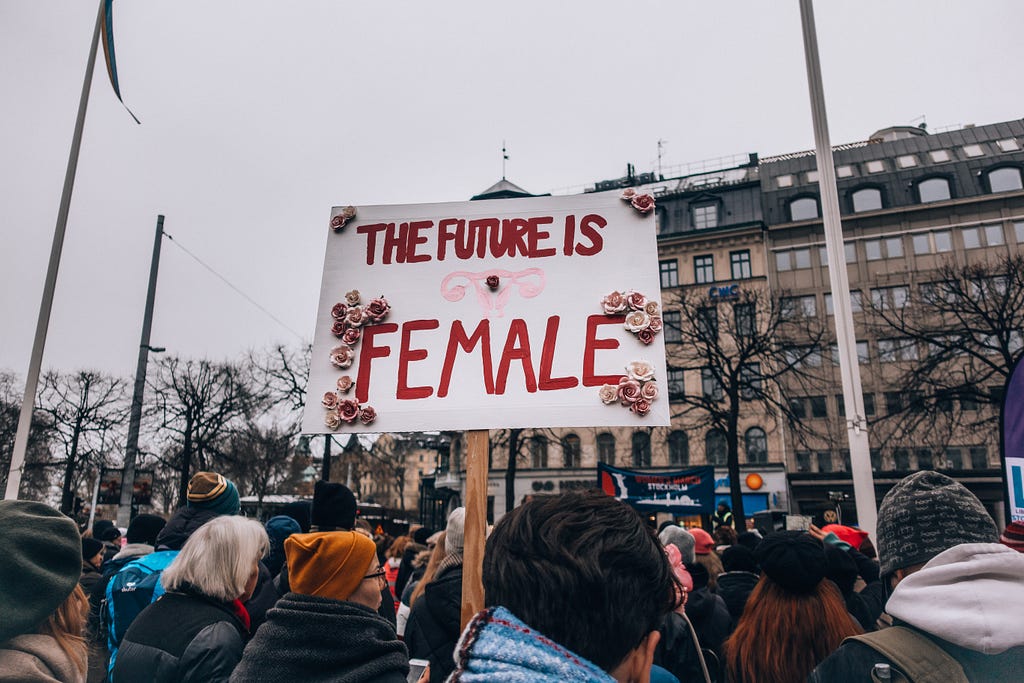 Someone in a crowd holds a sign that reads “The future is female” in large, bold, red letters. The photos is taken from behind the crowd, and the tops of peoples heads are visible, though most of them are wearing hats or beanies. A large building looms in the background.