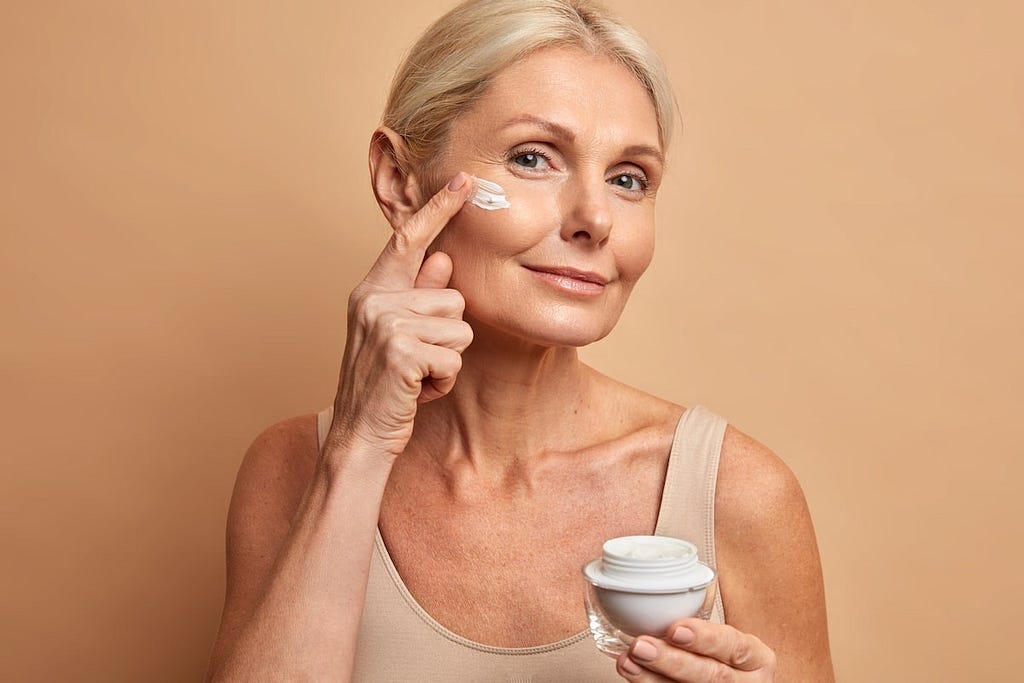 An image of a woman with blonde, graying hair smiling at the camera in front of cream background. In her right hand she holds an uncapped container of cream, while left applies some cream on her face.