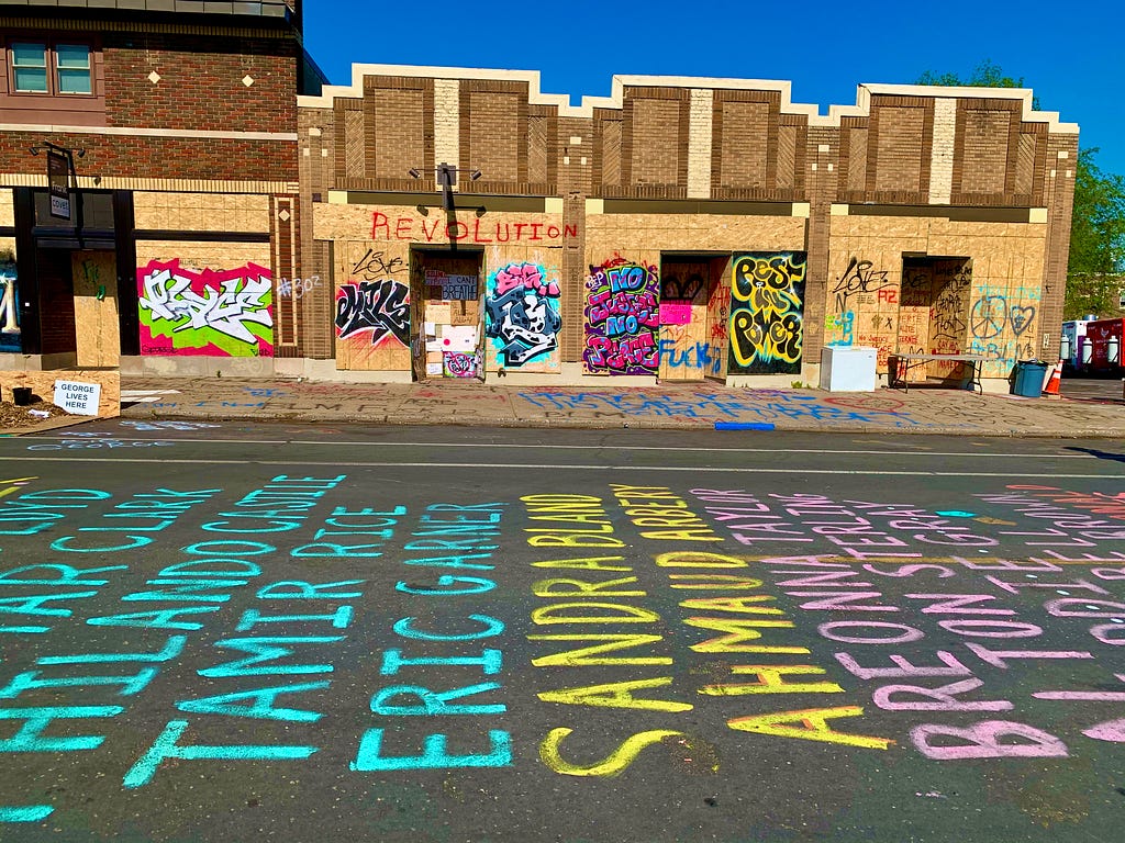 Boarded up storefronts with grafitti of social slogans such as “revolution” “rest in power” above a street with chalk list of the many victims of police violence Philando Castillo, Tamir Rice, Eric Garner, Sandra Bland, Ahmad Aurbery, Breonna Taylor, Alton Sterling, and more